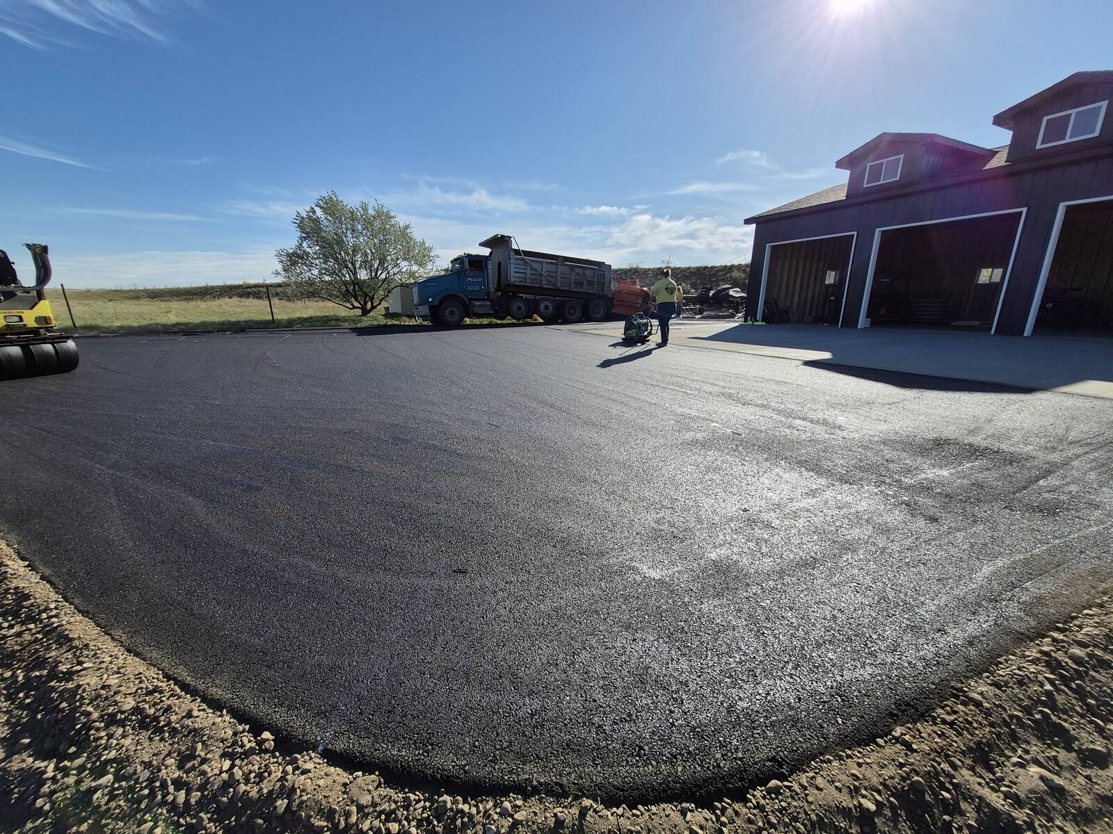 Wide new asphalt pad in front of a dark shop building with the paving crew finishing up
